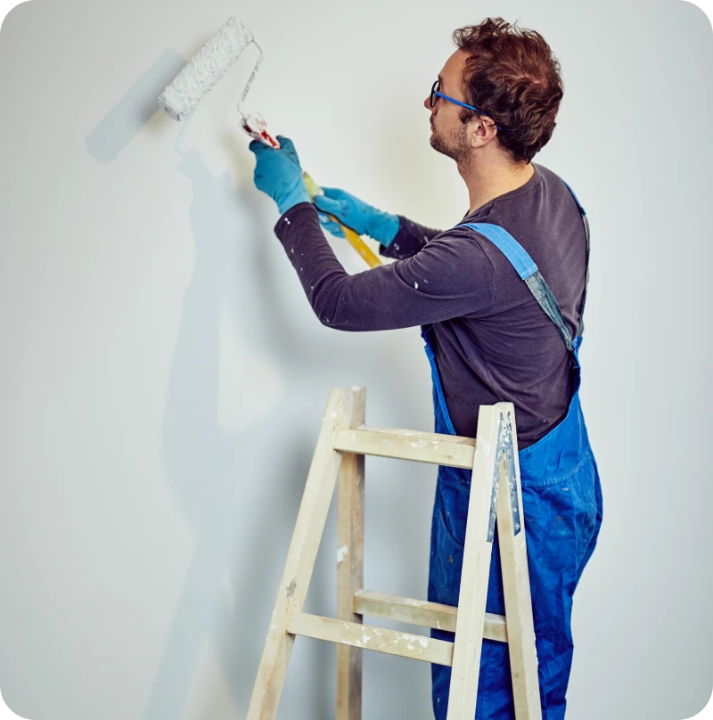 Person in overalls using paint roller indoors.